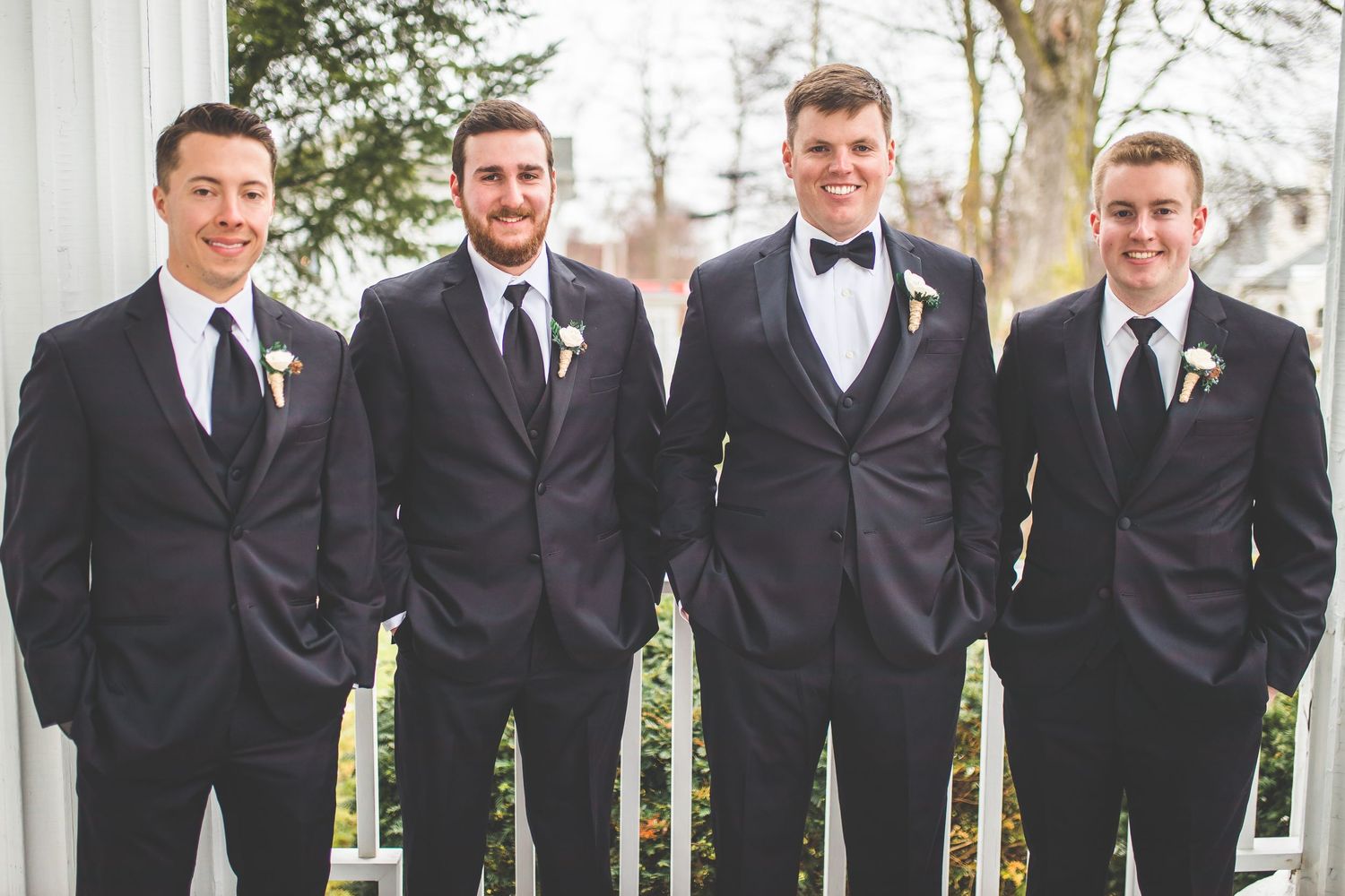 Groomsmen in black tuxedos stand in line against white columns outdoors.