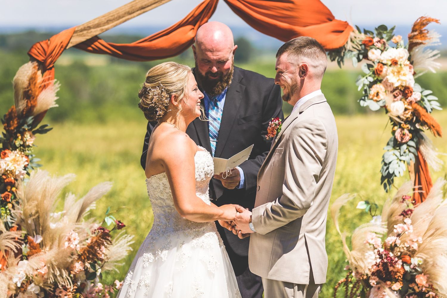 Wedding ceremony with orange fabric arch and floral arrangements in summer meadow.