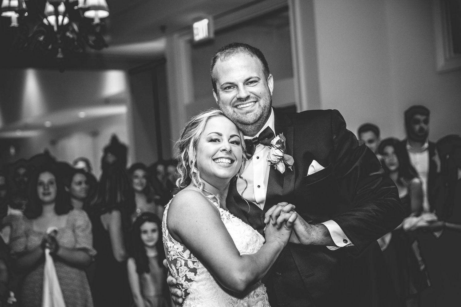 Black and white photo of a couple dancing at their wedding reception with guests in background.