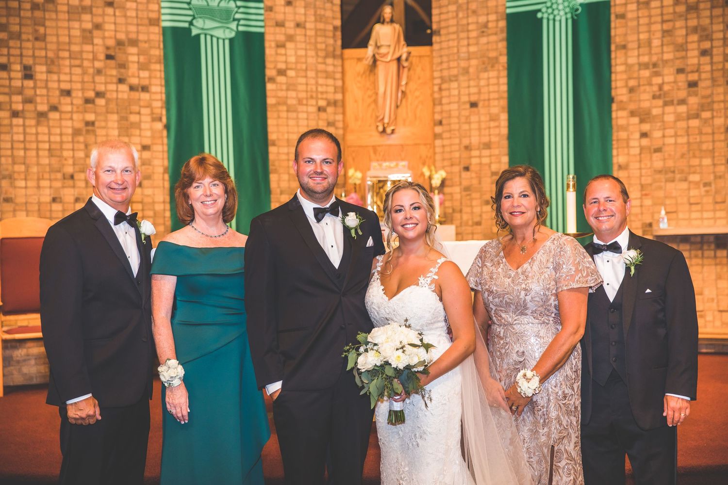 Wedding party poses for formal photos in front of brick church with green architectural details.