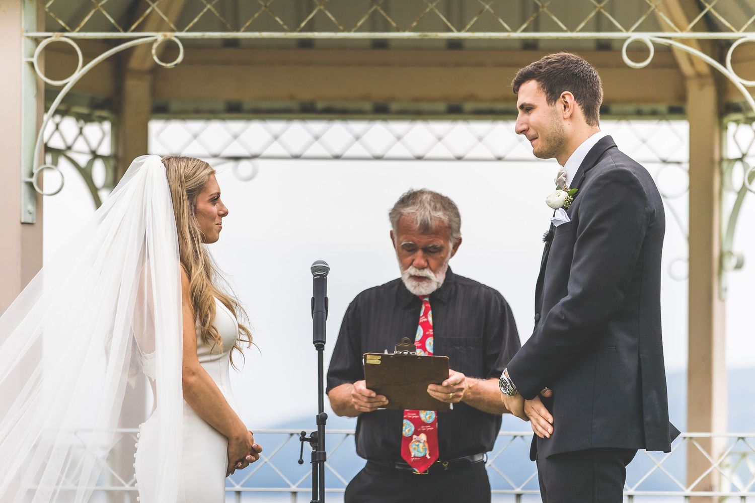 Wedding ceremony taking place under an ornate white gazebo overlooking scenic mountains and water.