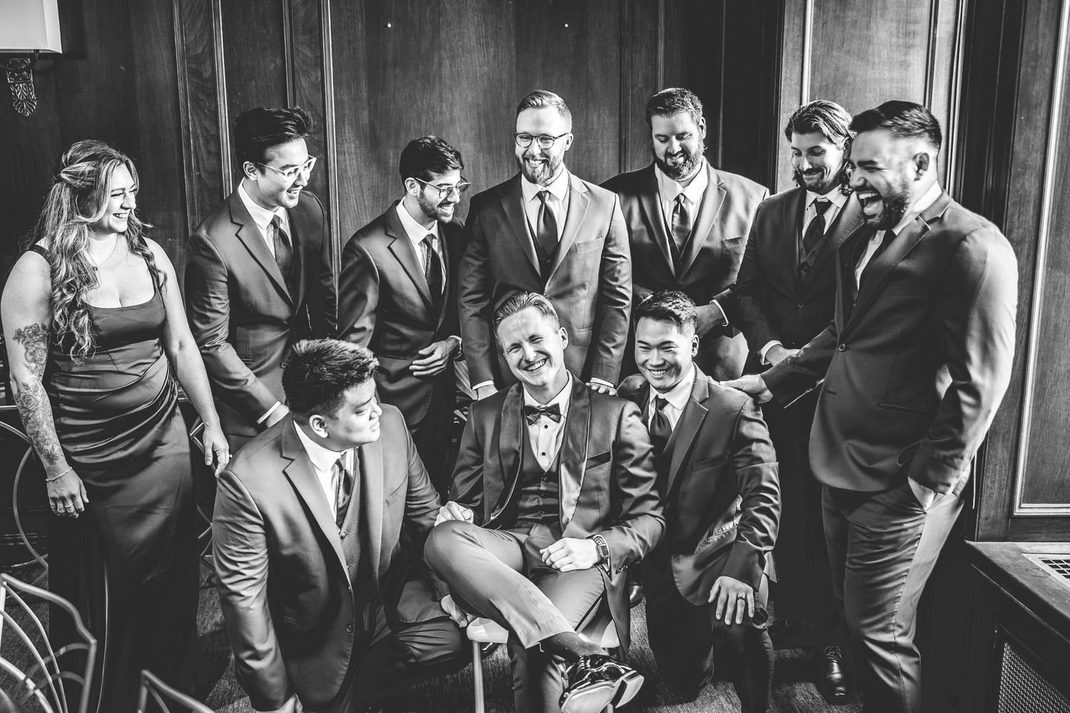 Black and white group photo of wedding party in formal attire gathered by window.
