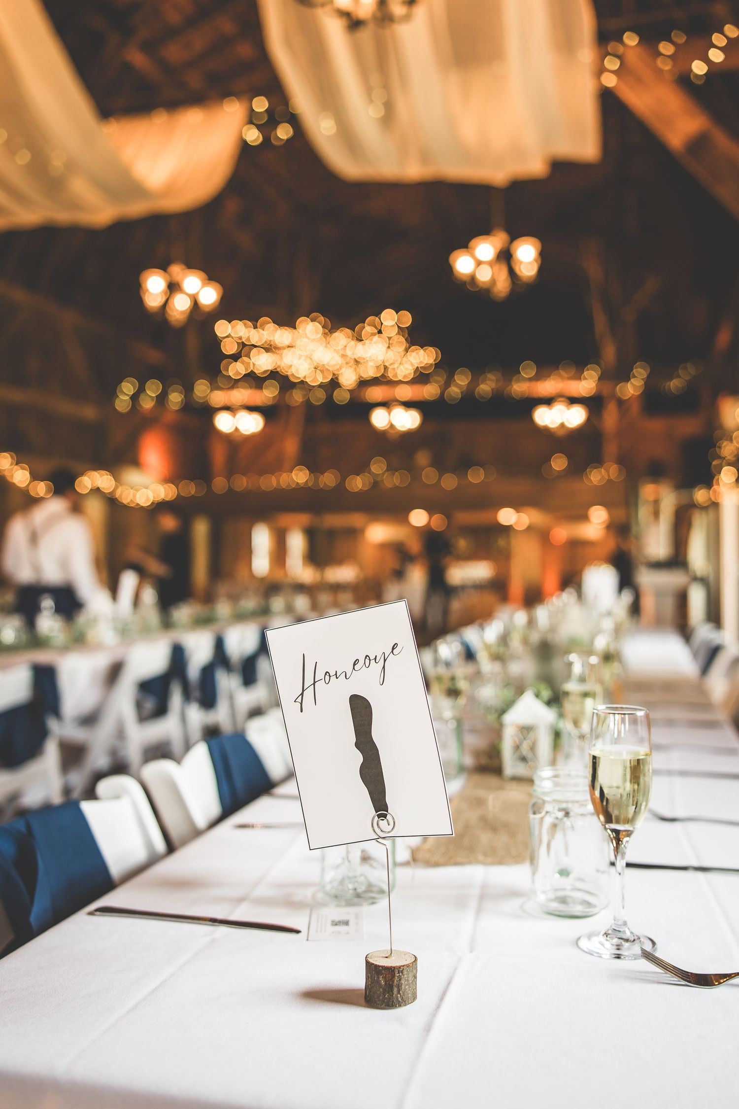 Wedding reception tables set with white linens and place cards in a rustic venue with warm lighting.