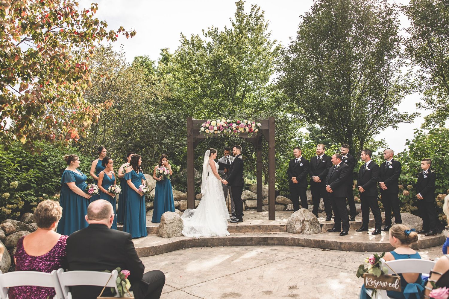 Outdoor wedding ceremony with guests seated on white chairs under trees and a wooden pergola.
