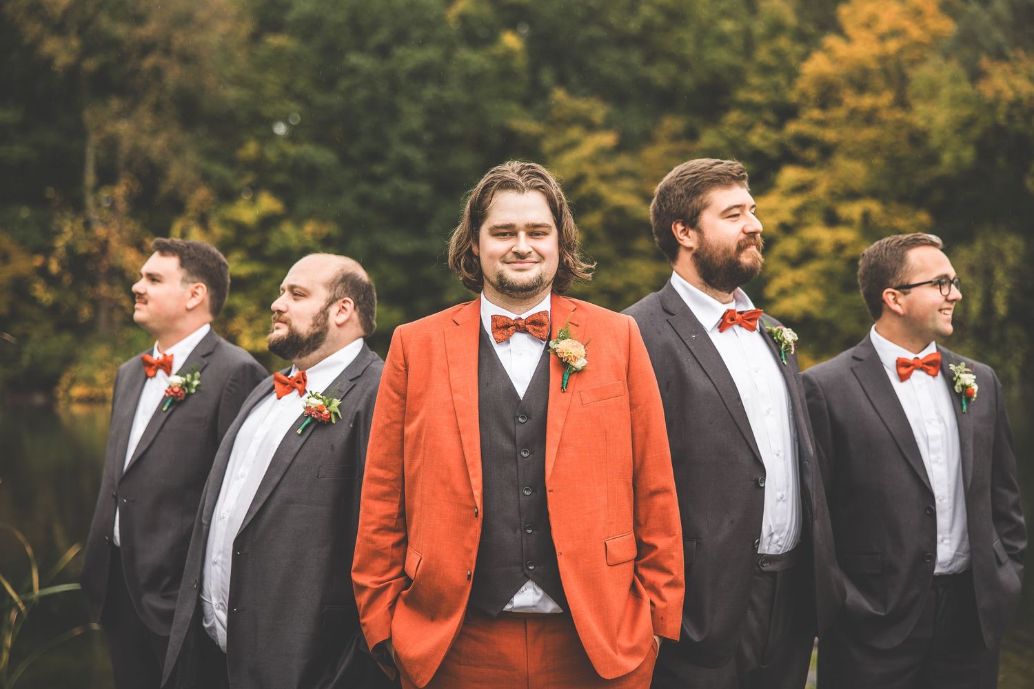 Groomsmen in black suits pose with one member in bright orange against an autumn lakeside backdrop.