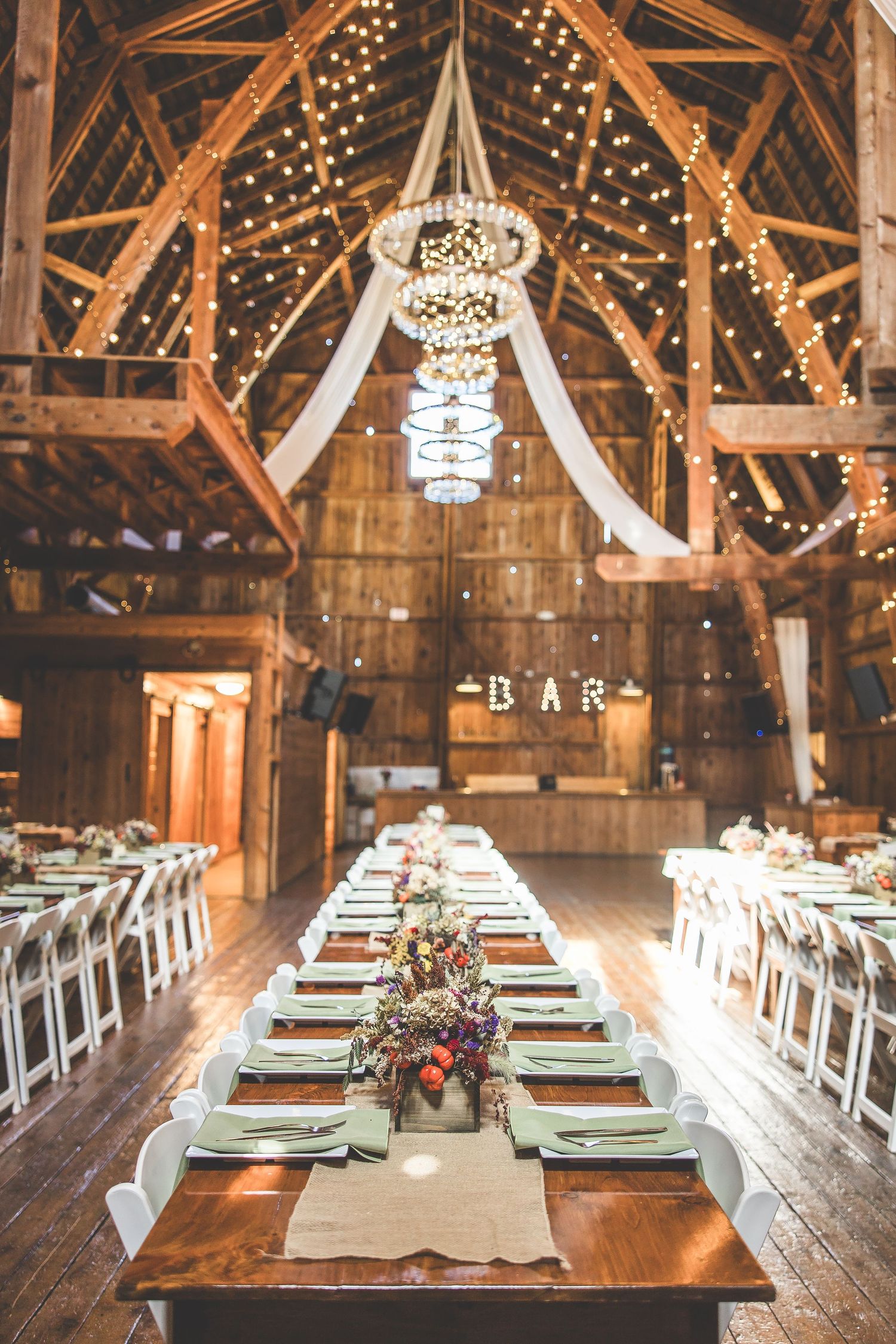 Rustic barn wedding reception setup with long wooden tables, white chairs, and a grand chandelier with string lights.