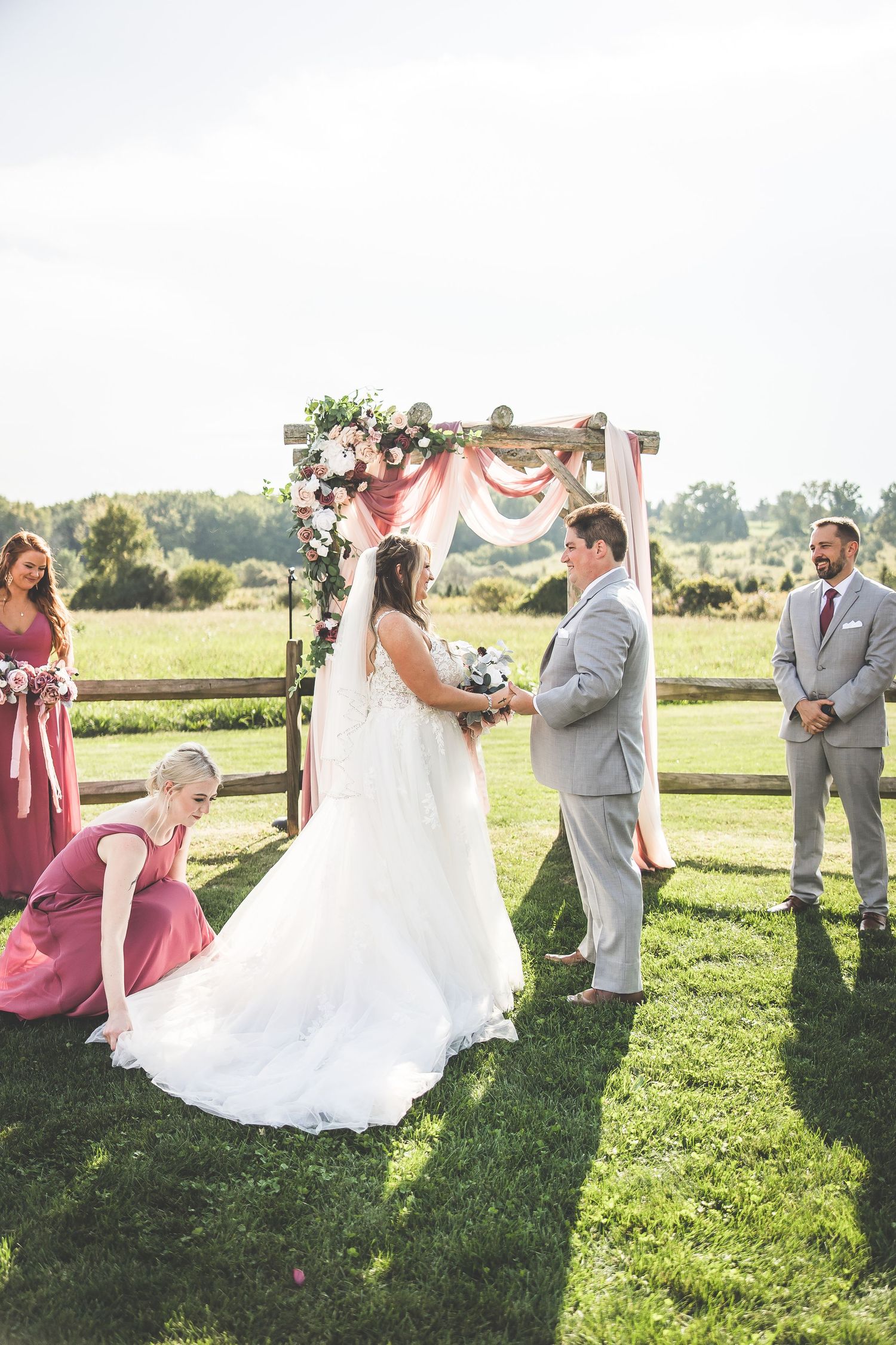 Outdoor wedding ceremony under pink and white floral arch with bridal party in mauve and grey attire.