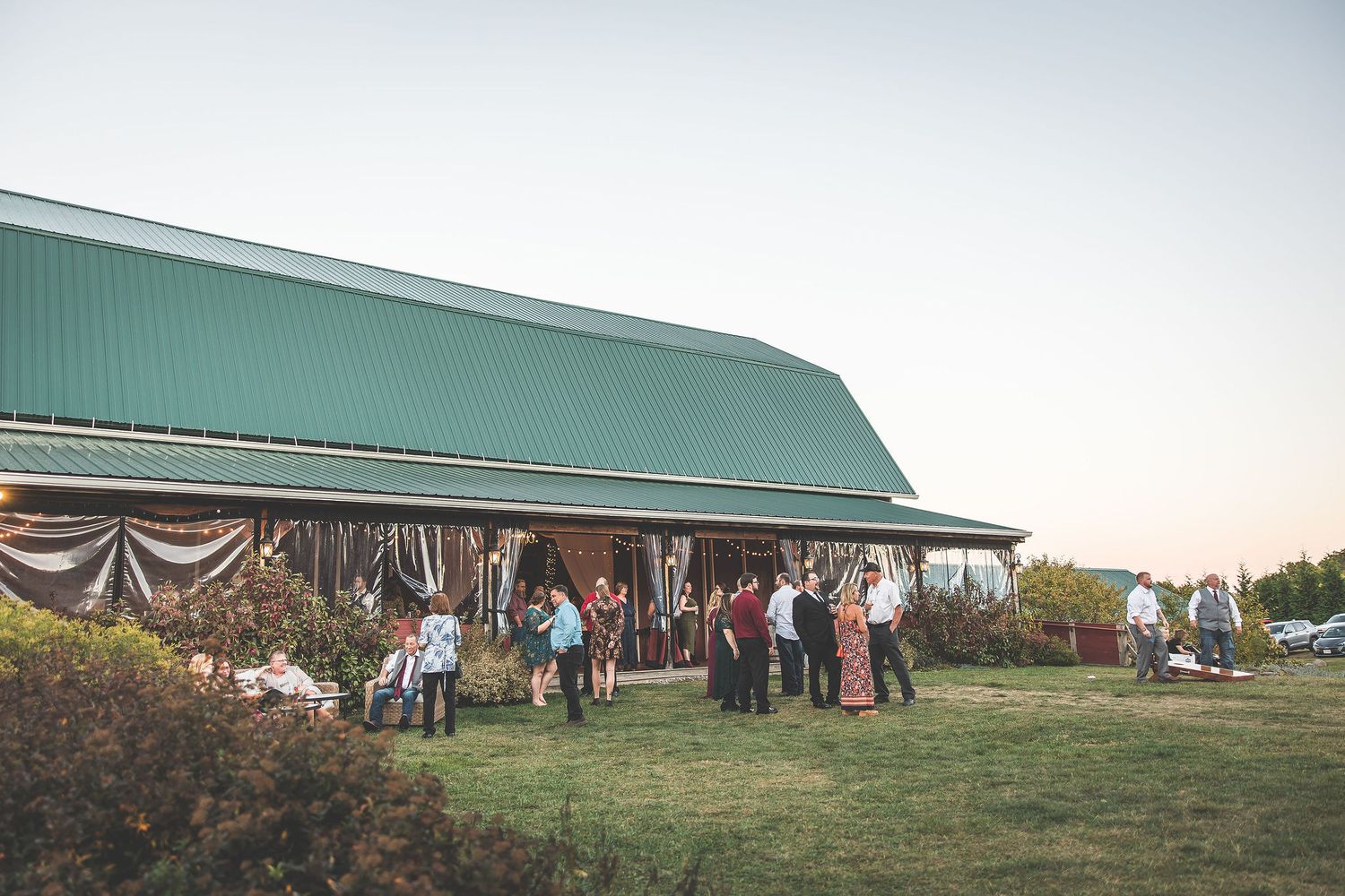 Wedding guests gather outside a green-roofed barn during an outdoor reception celebration.