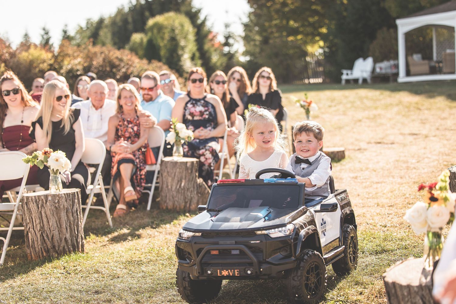 Outdoor wedding processional with toy cars and guests seated on hillside.