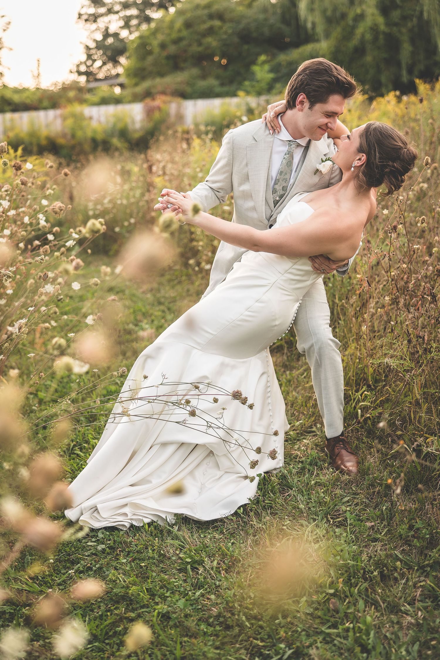 A romantic wedding couple dances in a wildflower field at sunset, with the bride's white dress flowing in the breeze.