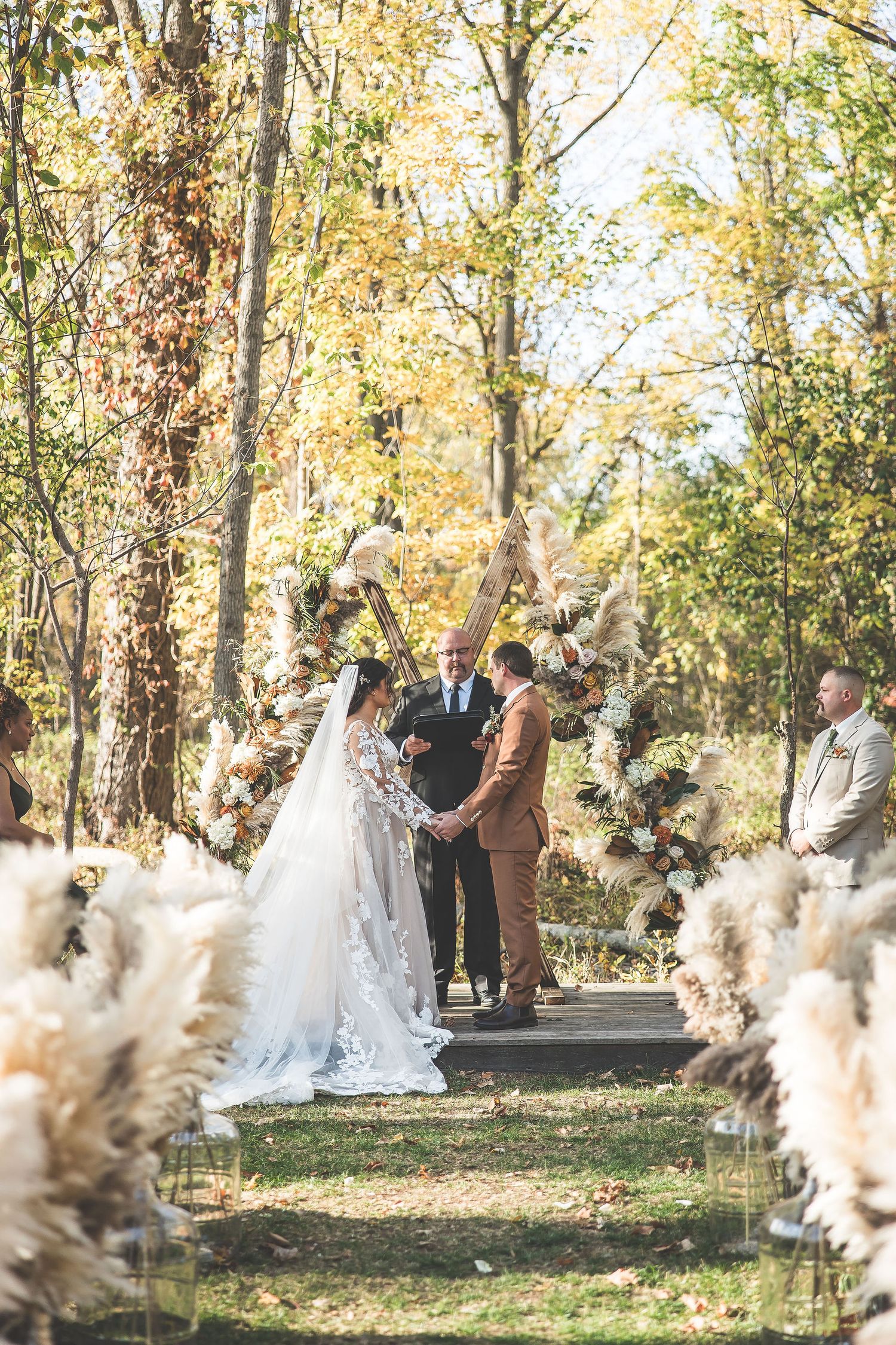 Autumn outdoor wedding ceremony with pampas grass decorations in a sunlit forest setting.