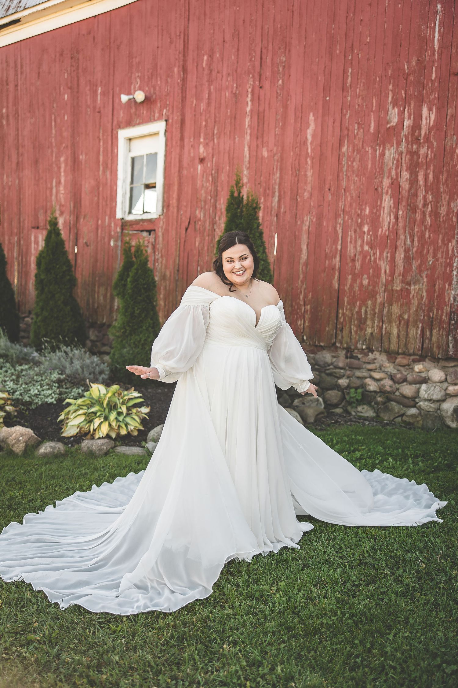A bride in a flowing white dress poses in front of a rustic red barn.