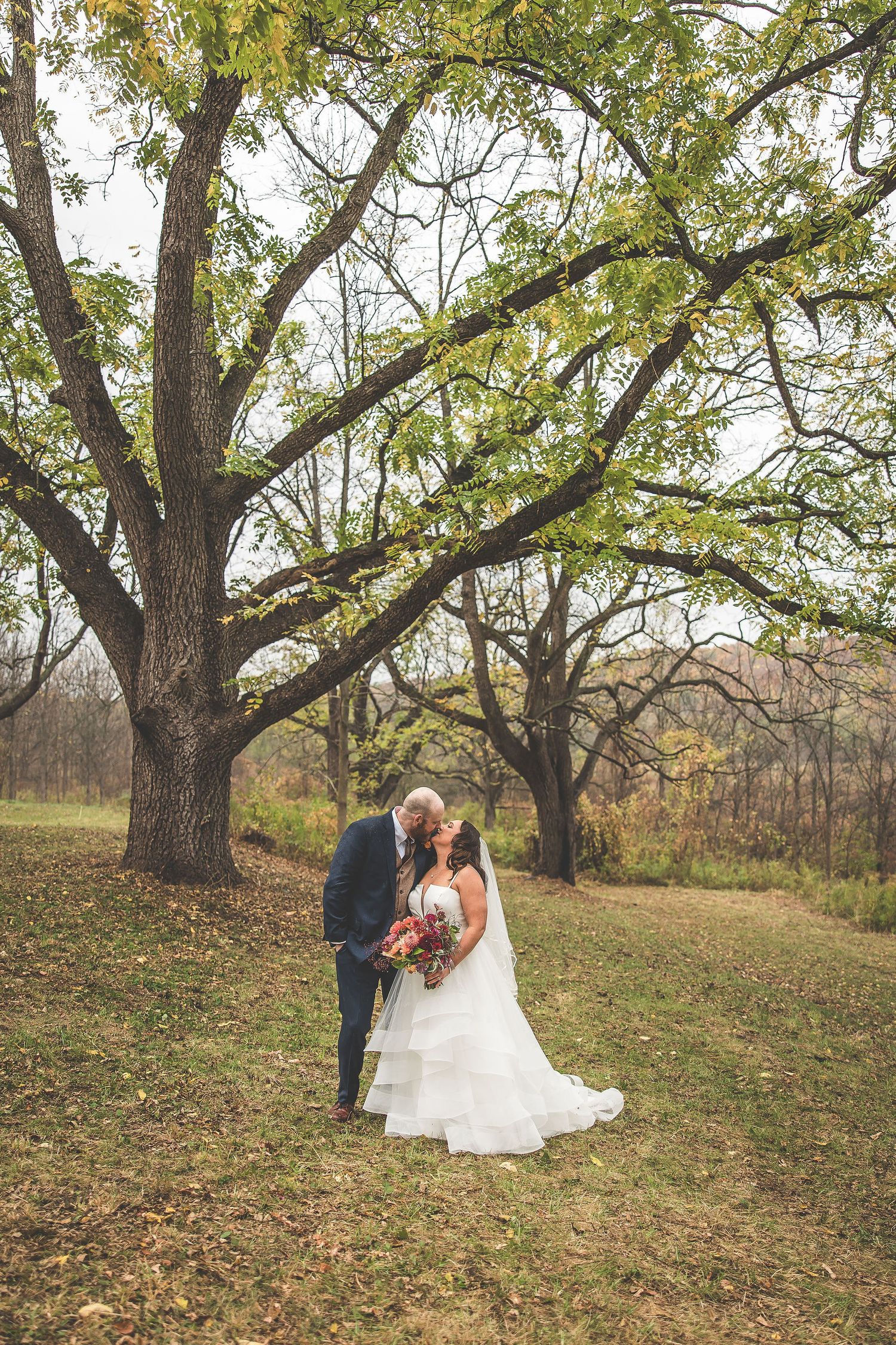 Newlyweds embrace during their fall wedding photoshoot among large trees with scattered leaves.