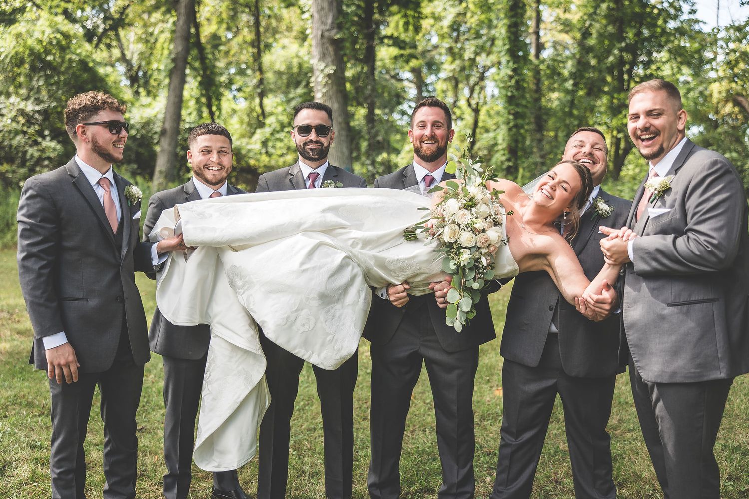 Groomsmen in grey suits pose playfully with the bride's dress train in a wooded setting.