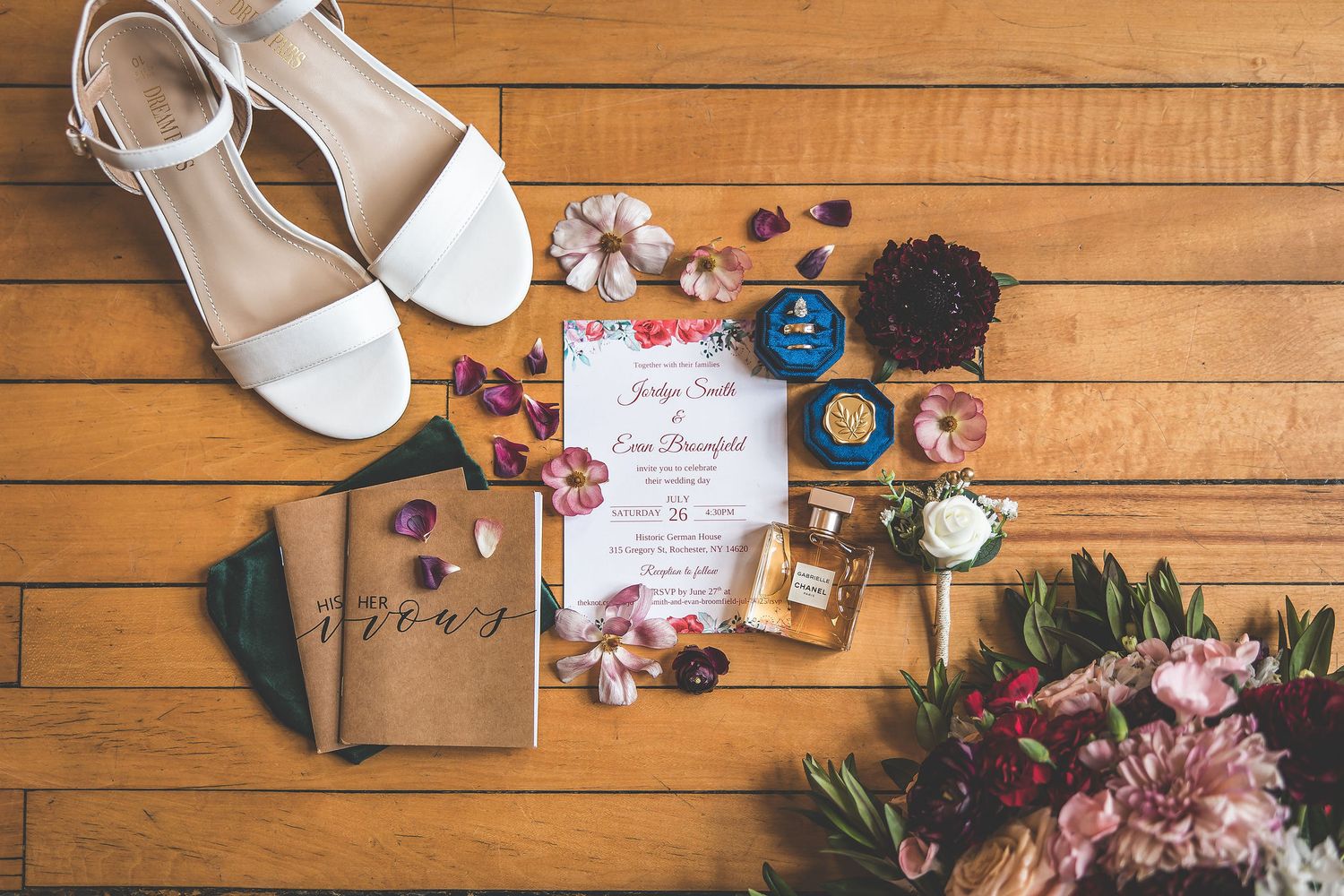 Wedding details flatlay with flowers, rings, and invitation on wooden surface.