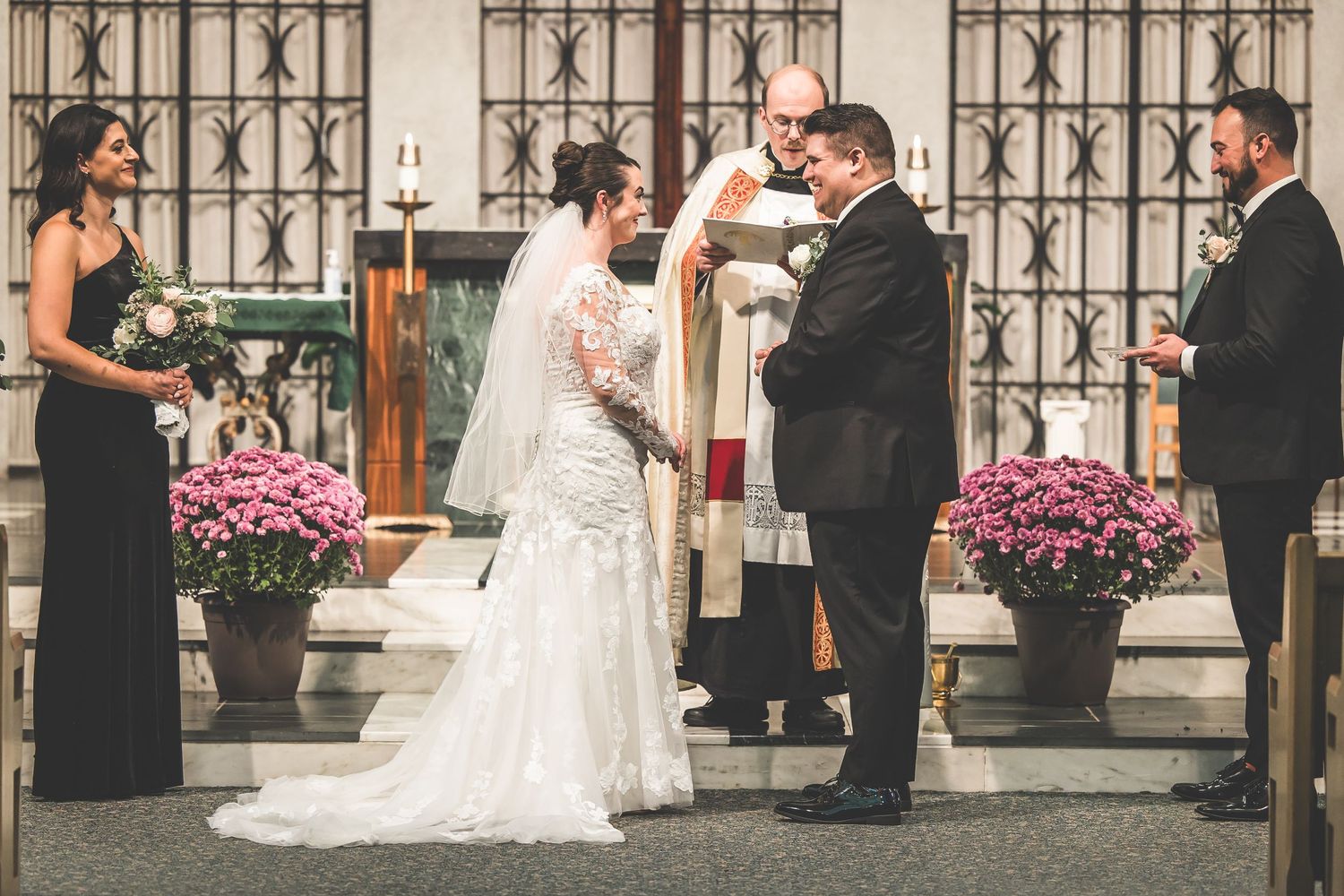 Wedding ceremony proceedings at church altar with decorative iron windows and pink flowers.