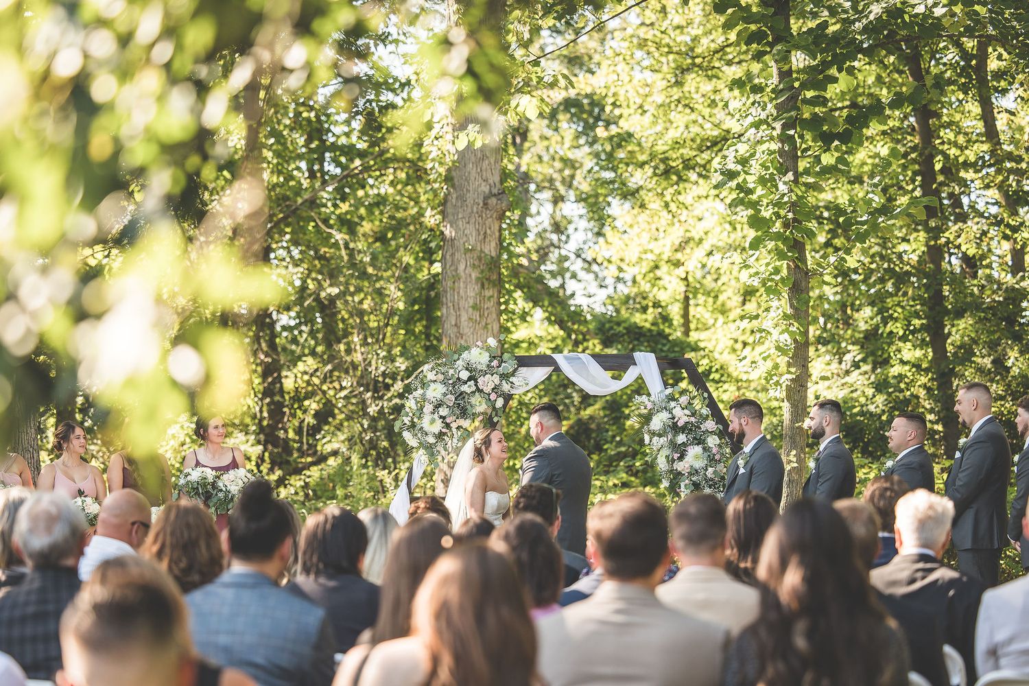 A wedding ceremony setup in a sunlit wooded area with rows of chairs and guests gathered.