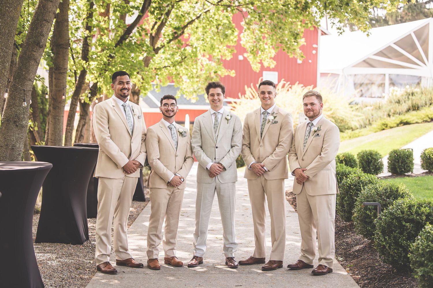 Wedding party in cream colored suits posed outdoors under green trees.