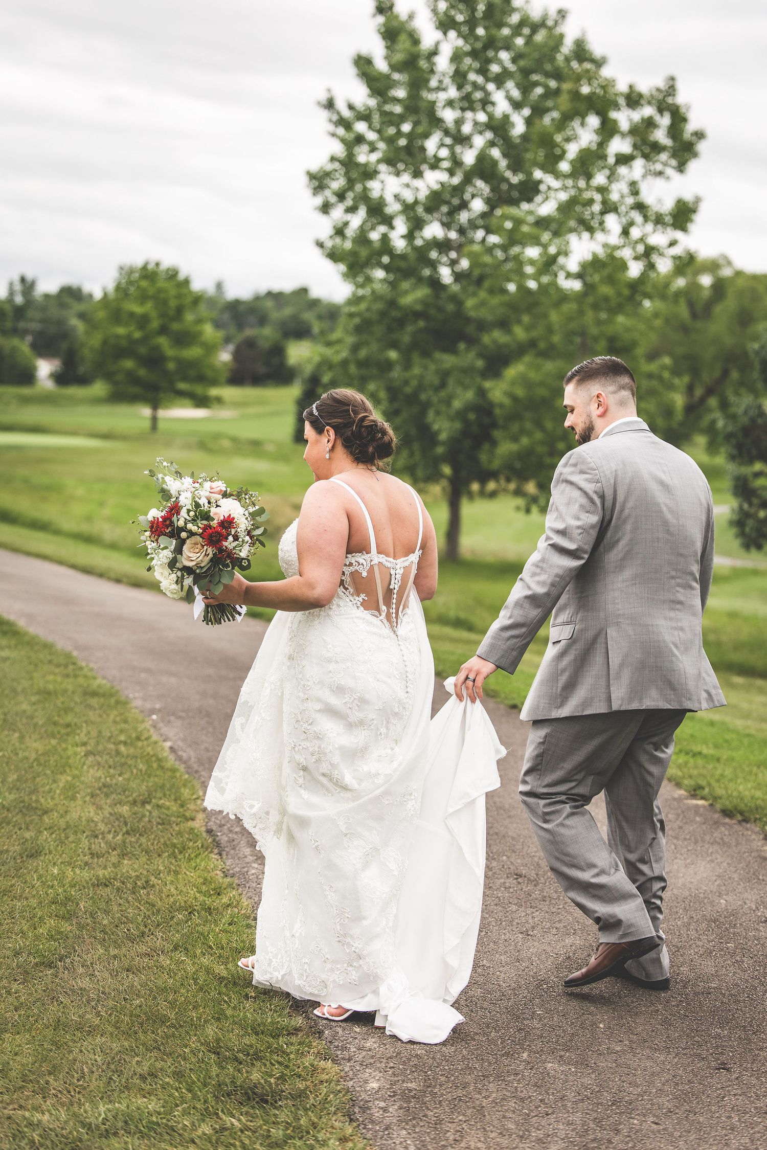 A couple walks down a path as the bride holds a vibrant bouquet and her white lace dress flows behind her.
