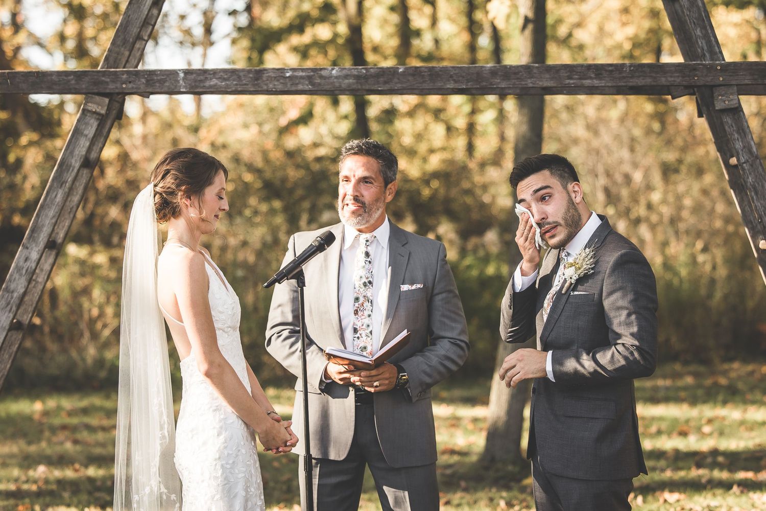 An outdoor wedding ceremony takes place under a wooden arch with autumn trees in the background.