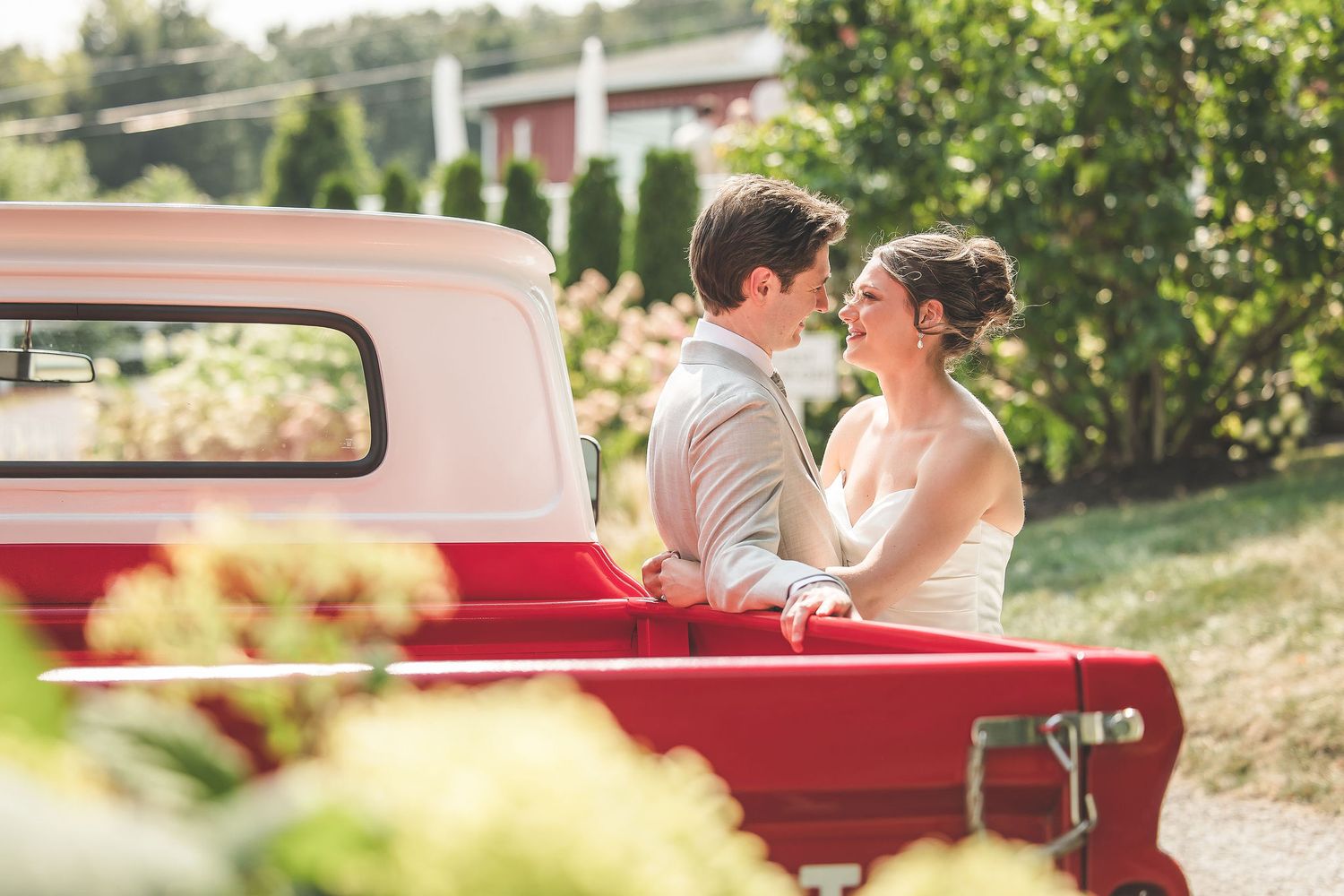 Tender moments captured with newlyweds and a vintage red pickup truck in sunlit garden setting.