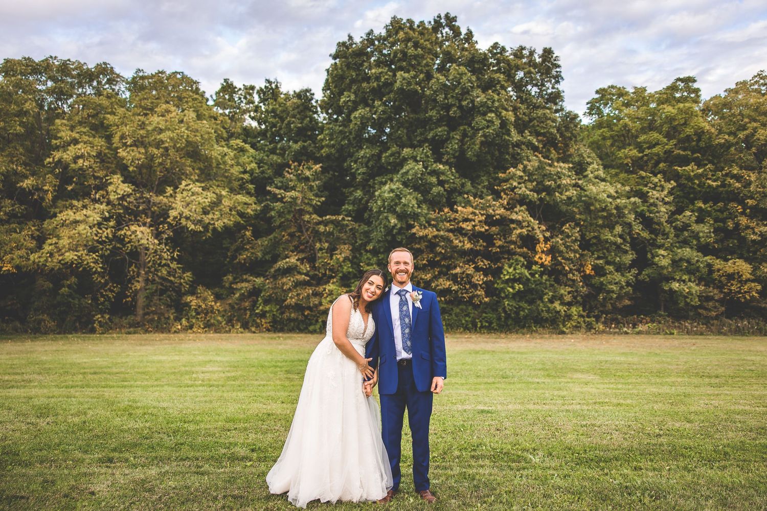 Wedding couple poses for romantic portraits in front of tree line on sprawling grass field.
