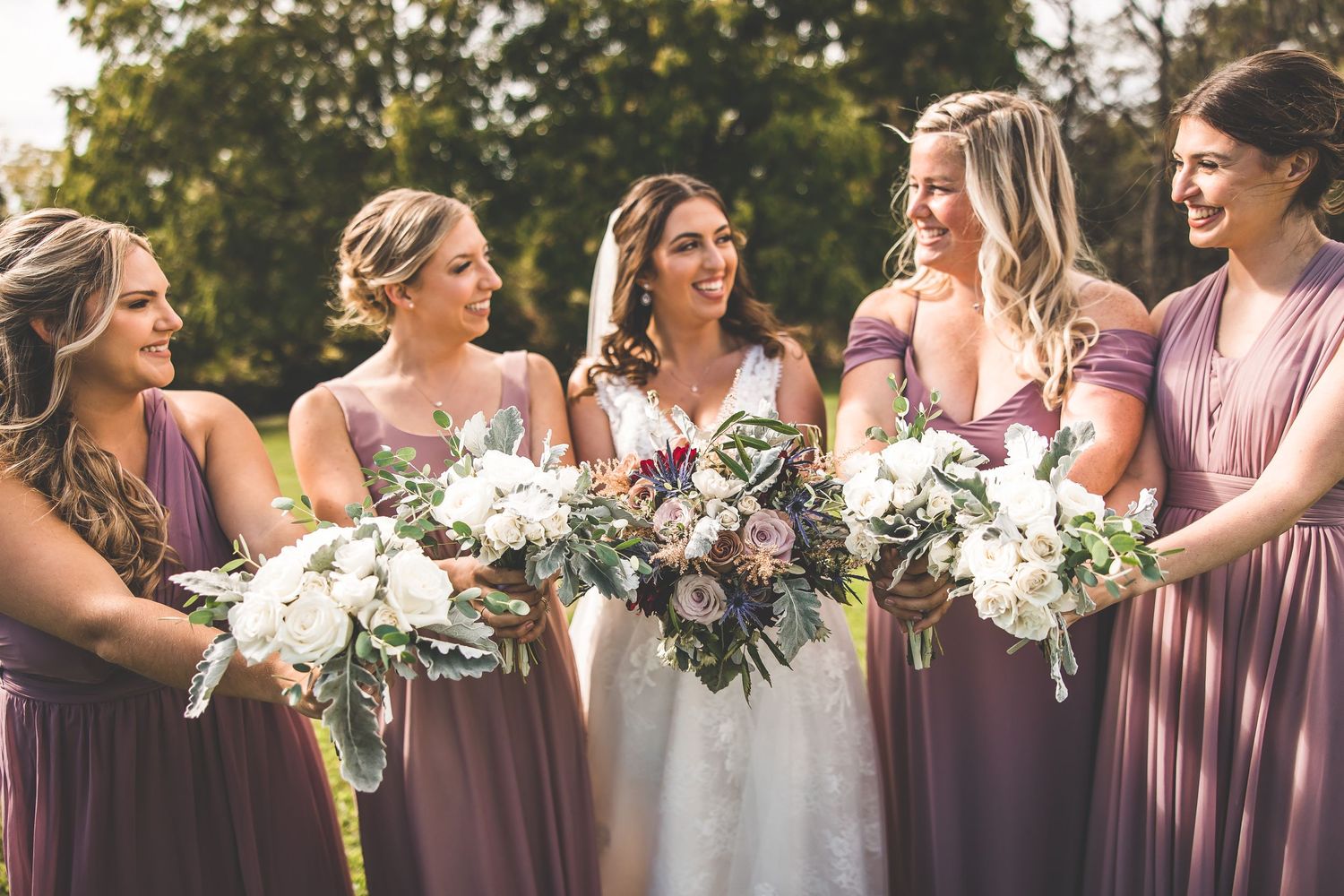 Bridesmaids in mauve dresses share candid moment while holding white floral bouquets outdoors.