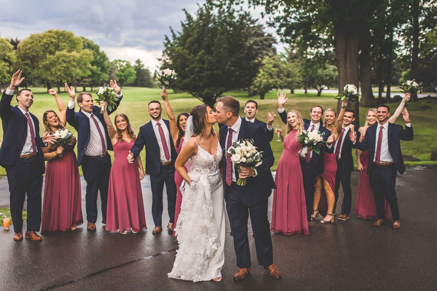 Large wedding party stands in line on grassy lawn with bridesmaids in burgundy and groomsmen in navy suits.