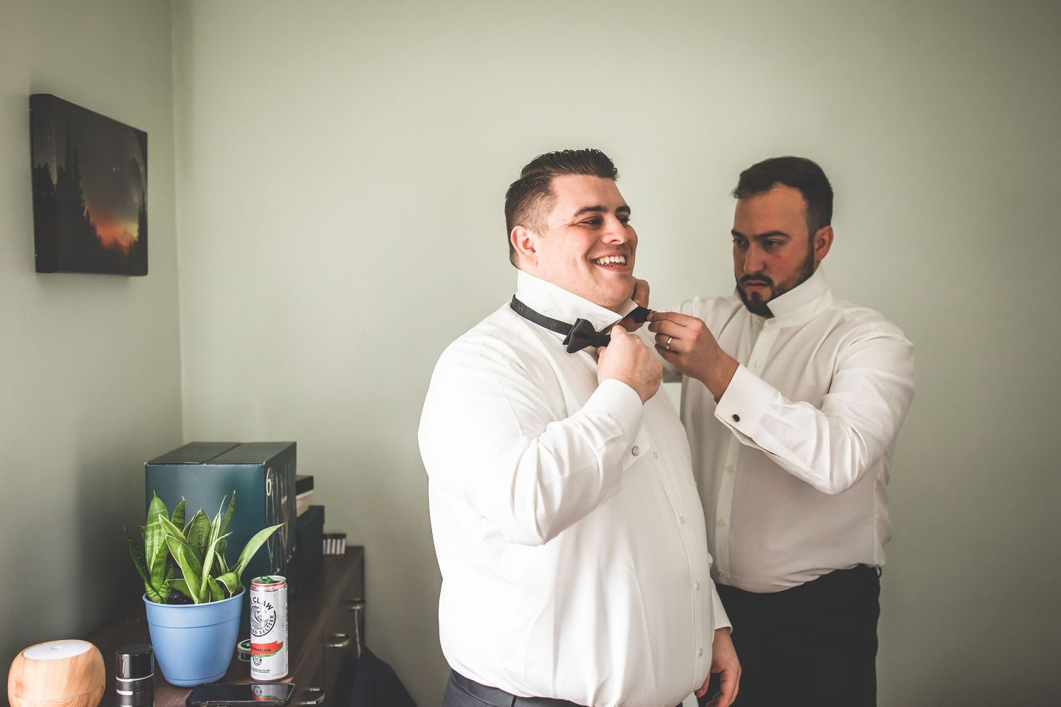 Two people in white dress shirts making final adjustments to formal attire in a room.