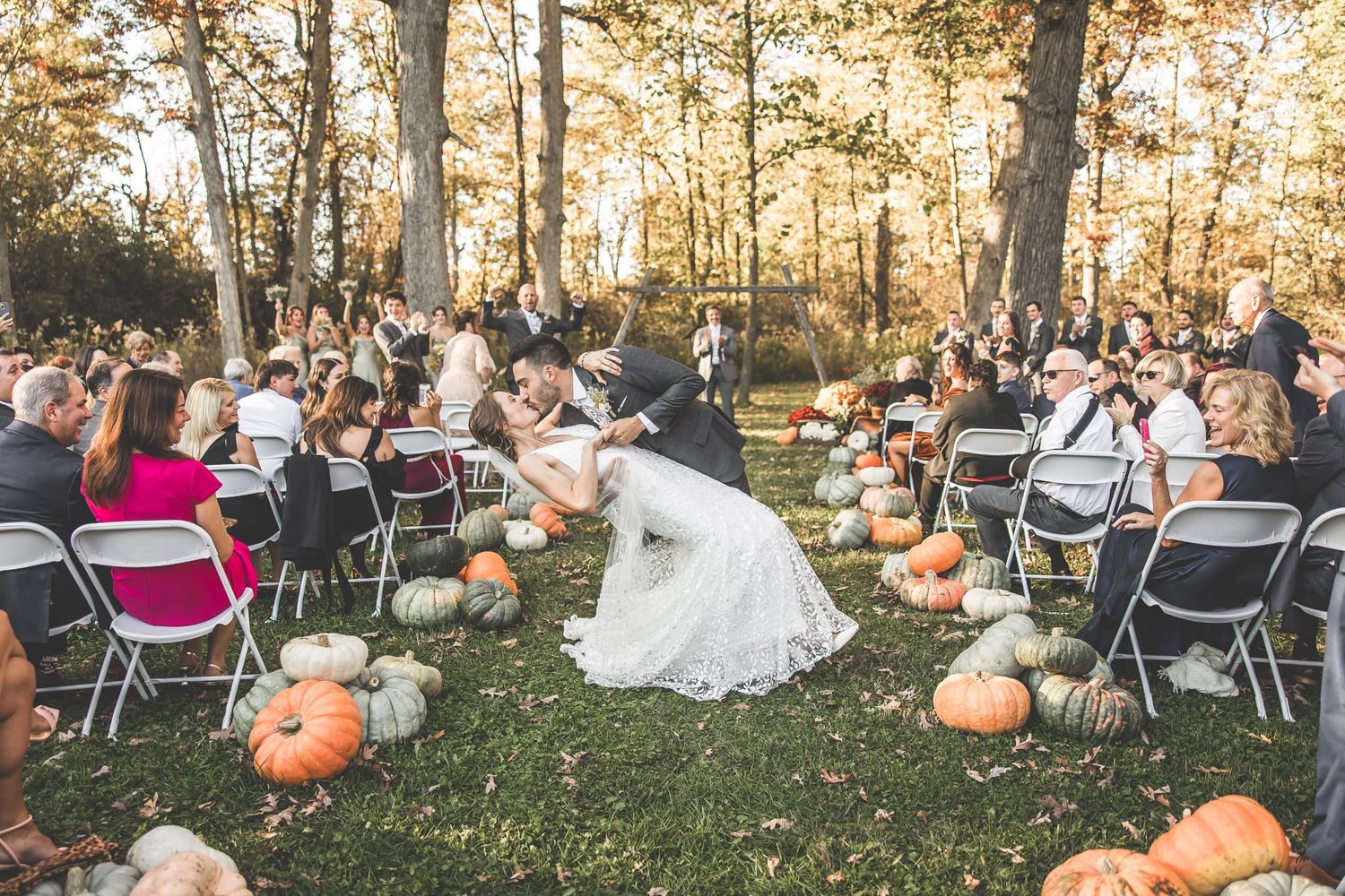 Wedding guests gathered in a wooded outdoor venue with scattered pumpkins and autumn decor.