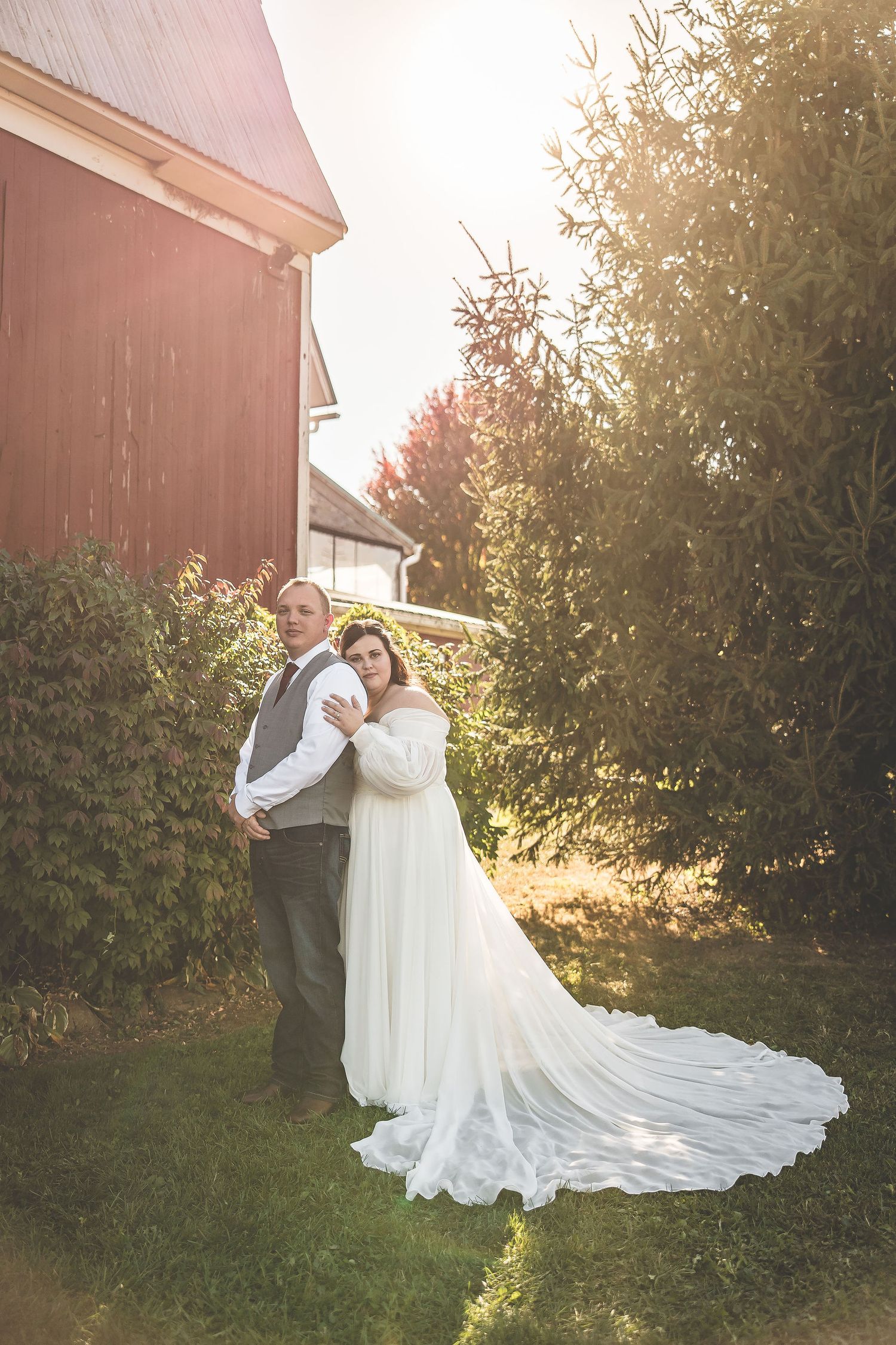 Wedding couple poses in golden sunlight next to a red barn with a flowing white dress train.
