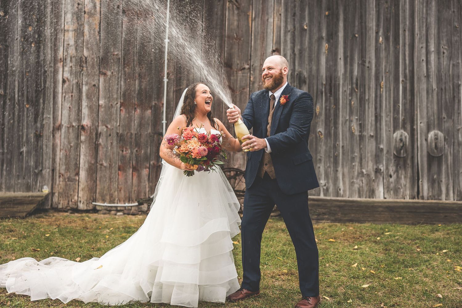 Wedding couple poses playfully in front of a rustic wooden barn backdrop.