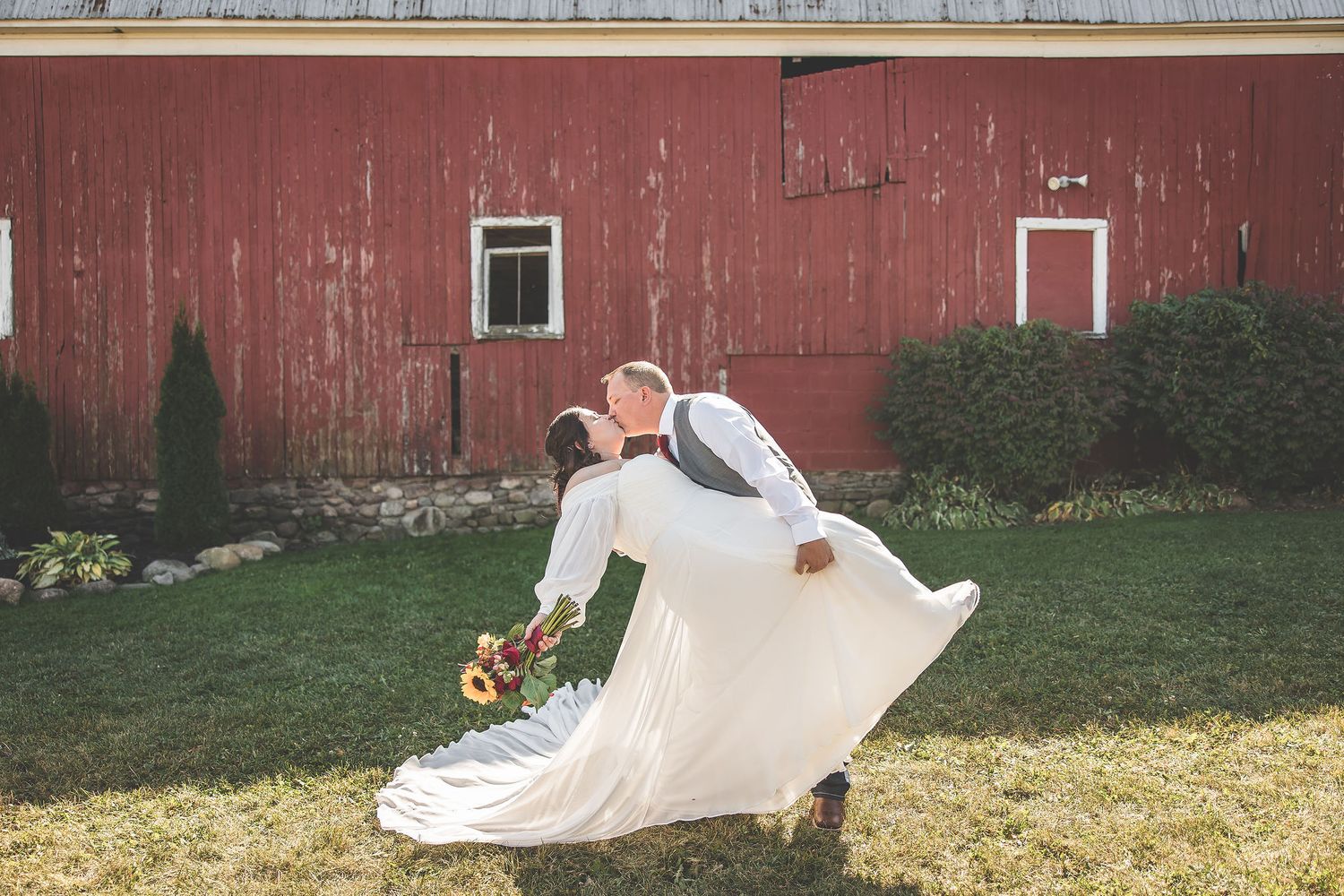 Bride's flowing dress train spreads across grass by rustic barn wall.