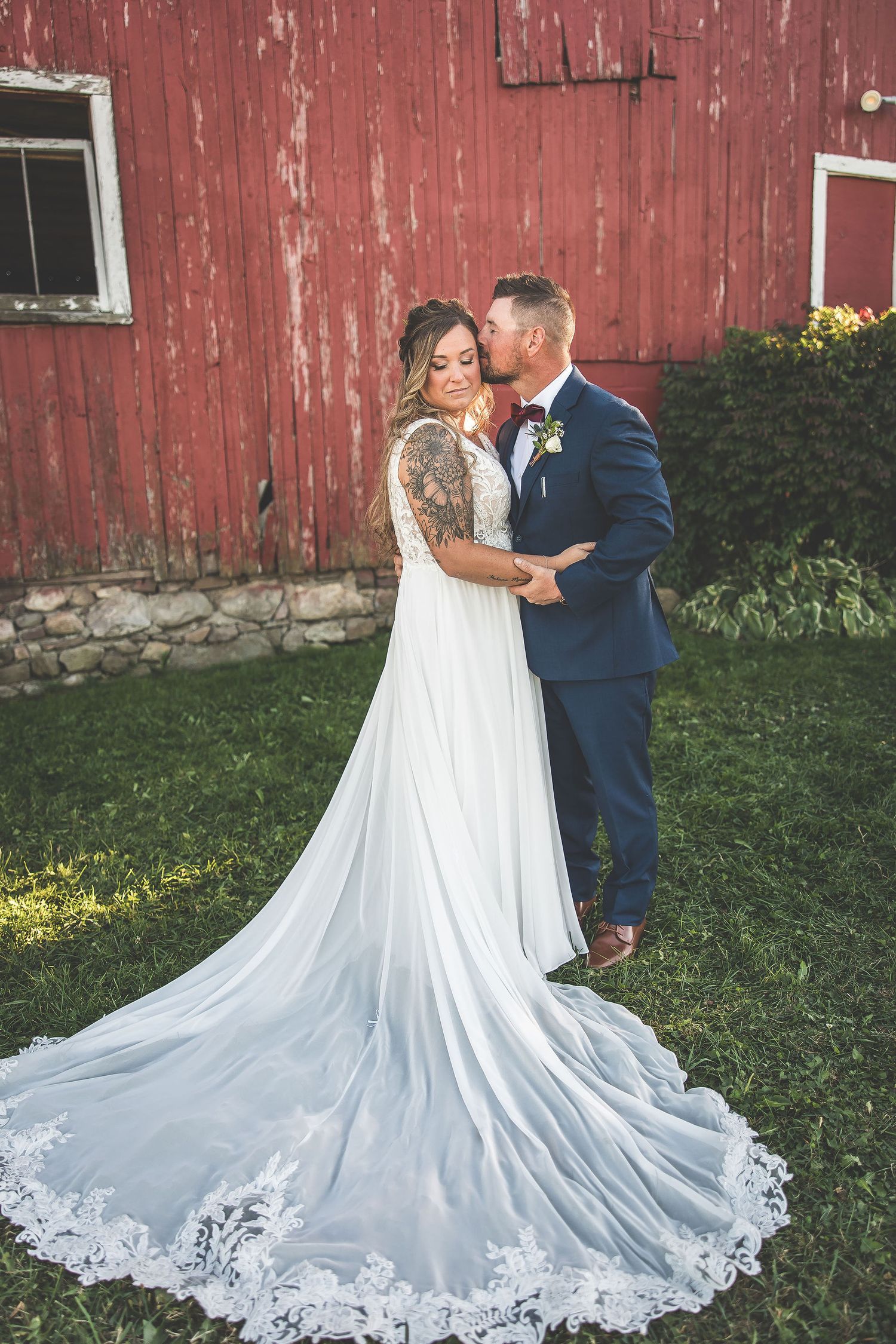 Wedding couple embraces by weathered red barn wall with dramatic white lace train spread on grass.