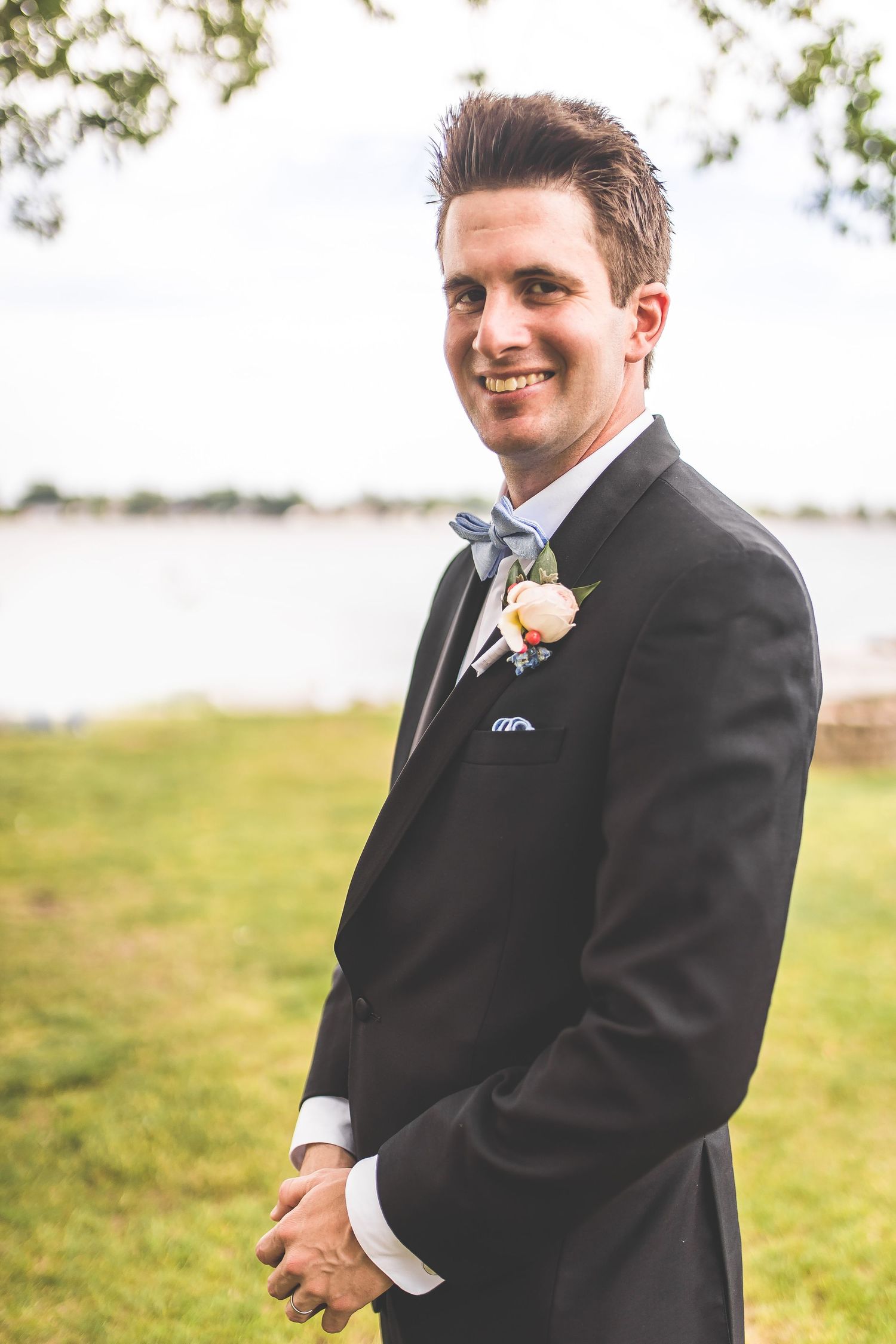 Groom in black suit with boutonniere poses outdoors on grassy lawn.