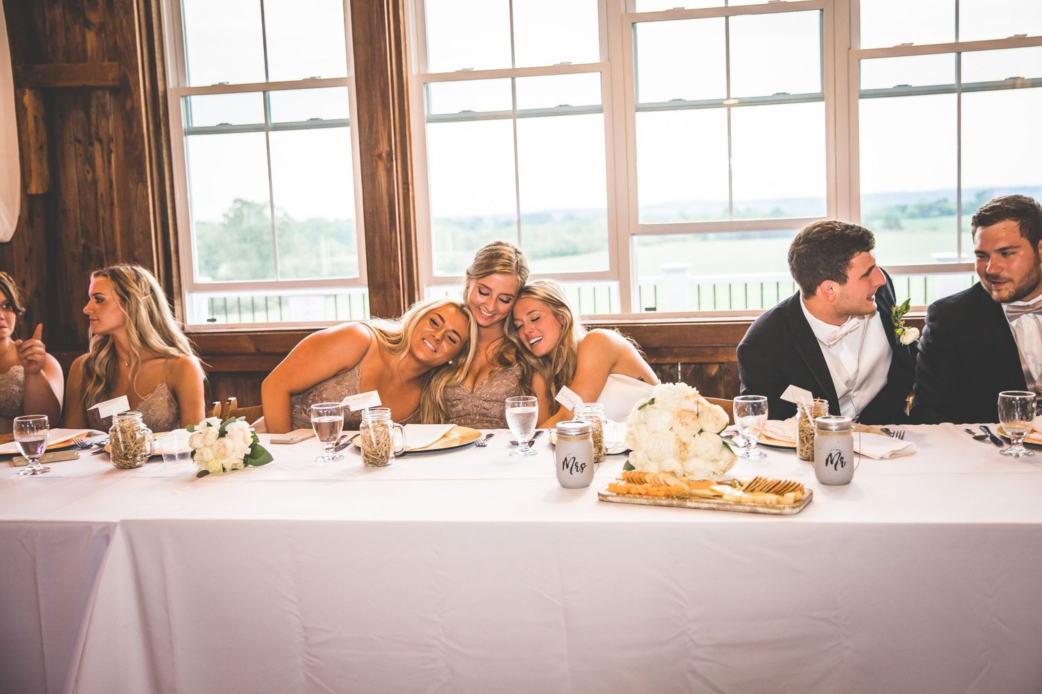 Wedding reception guests seated at long wooden tables in a rustic venue with large windows.