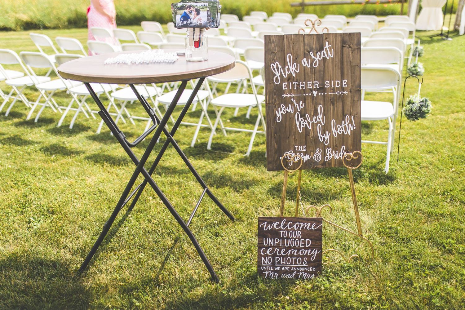 White folding chairs and chalkboard signs set up for an outdoor wedding ceremony.