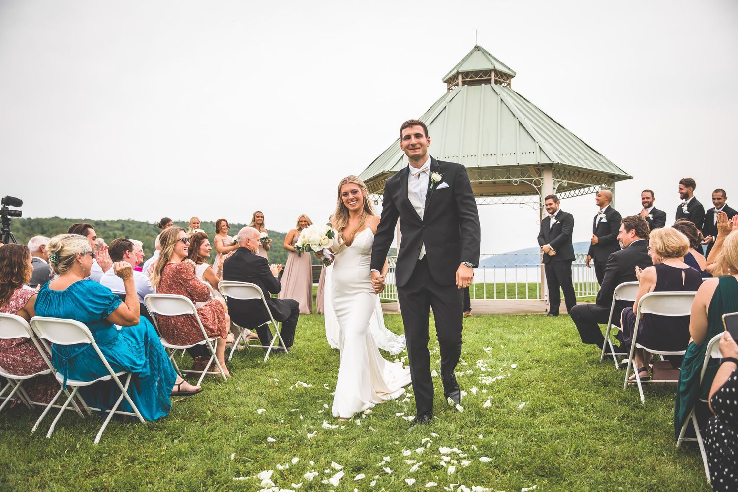 Wedding guests seated in white chairs on grass lawn with gazebo and lake view.