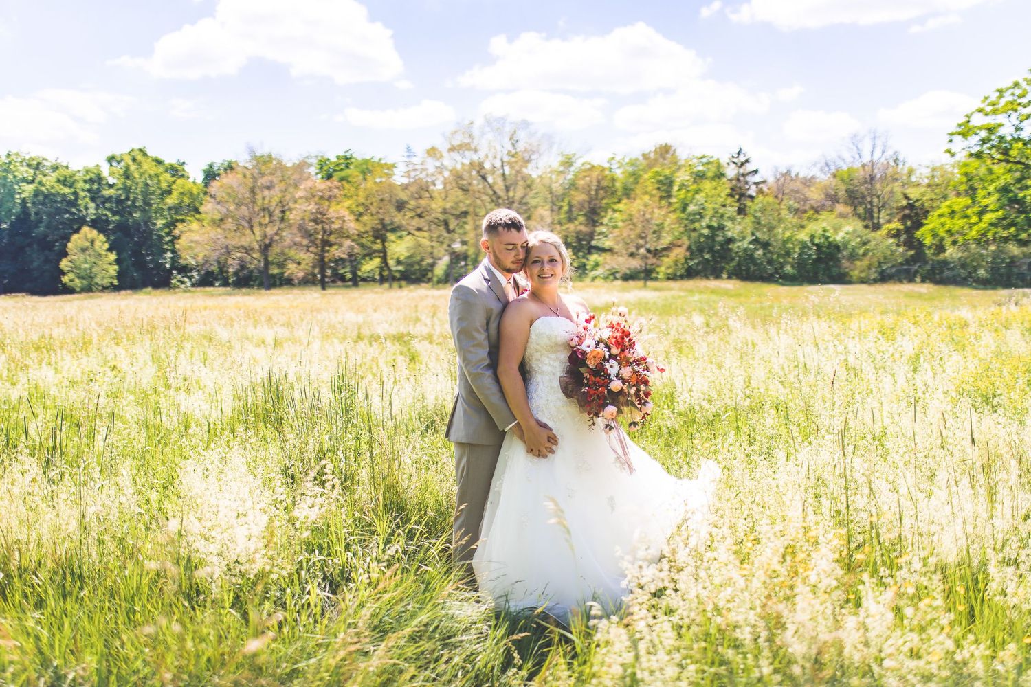 Wedding couple photographed in a serene meadow landscape with blue sky and wispy clouds.