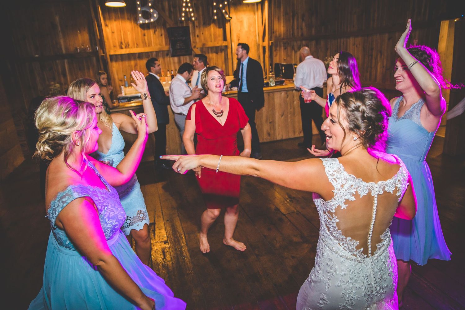 Wedding guests dancing and celebrating on a rustic wooden dance floor with colorful lighting.