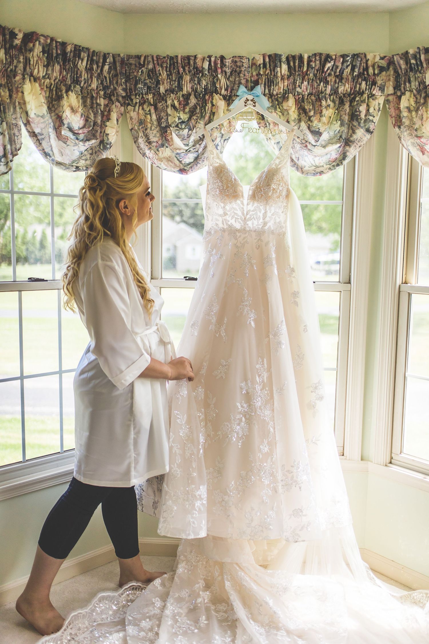 A beautiful white lace wedding dress hangs in a sunlit room with floral curtains.
