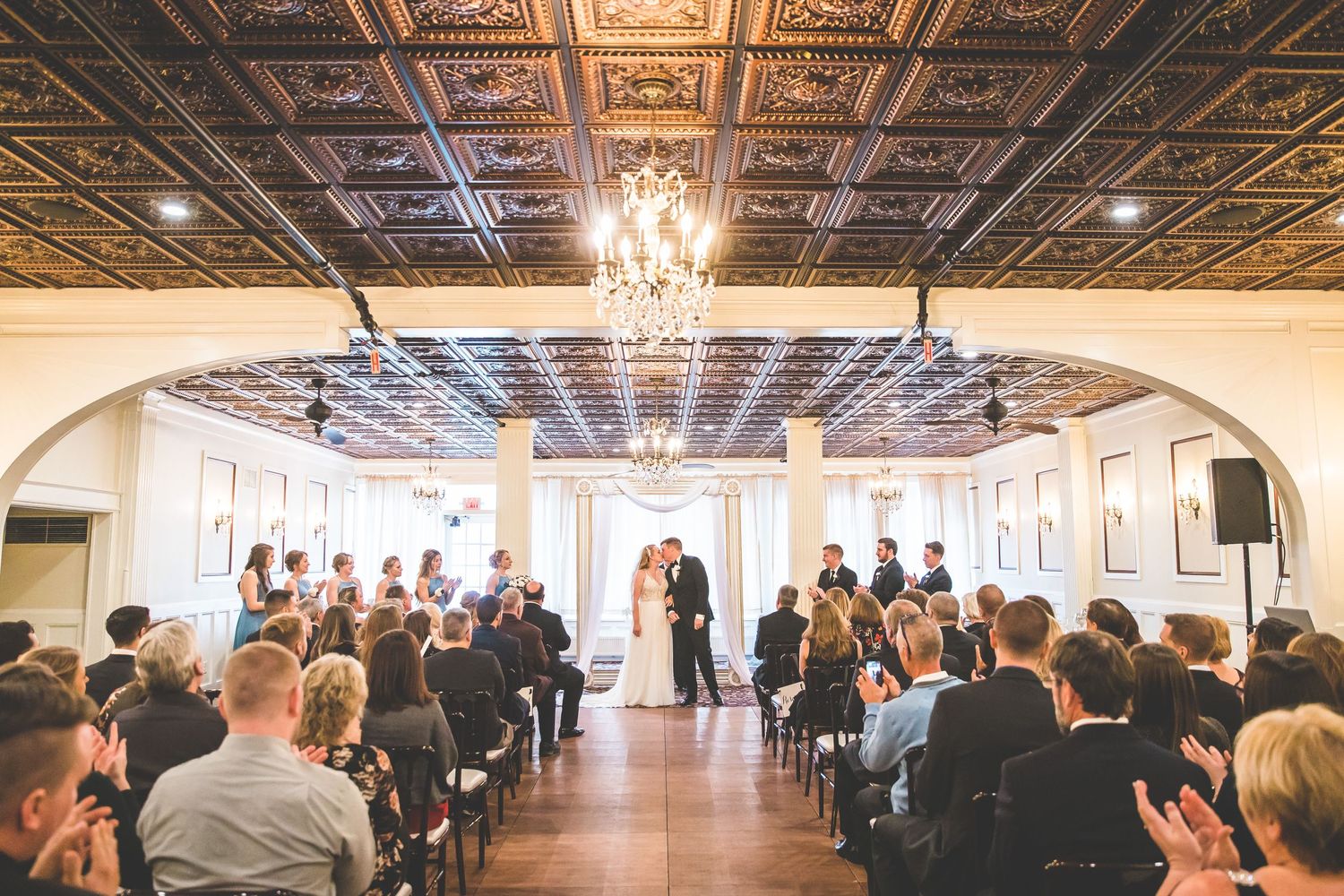 Elegant ballroom with ornate ceiling, chandeliers and white columns set up for a formal wedding ceremony.