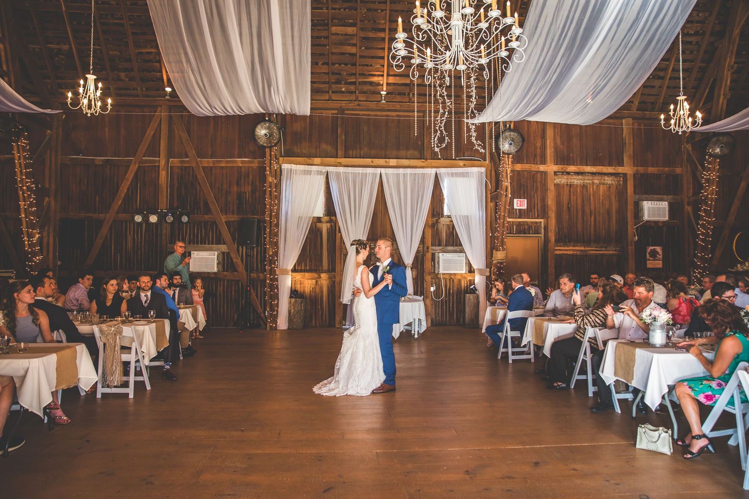 Rustic barn wedding reception venue with white draping and crystal chandeliers.
