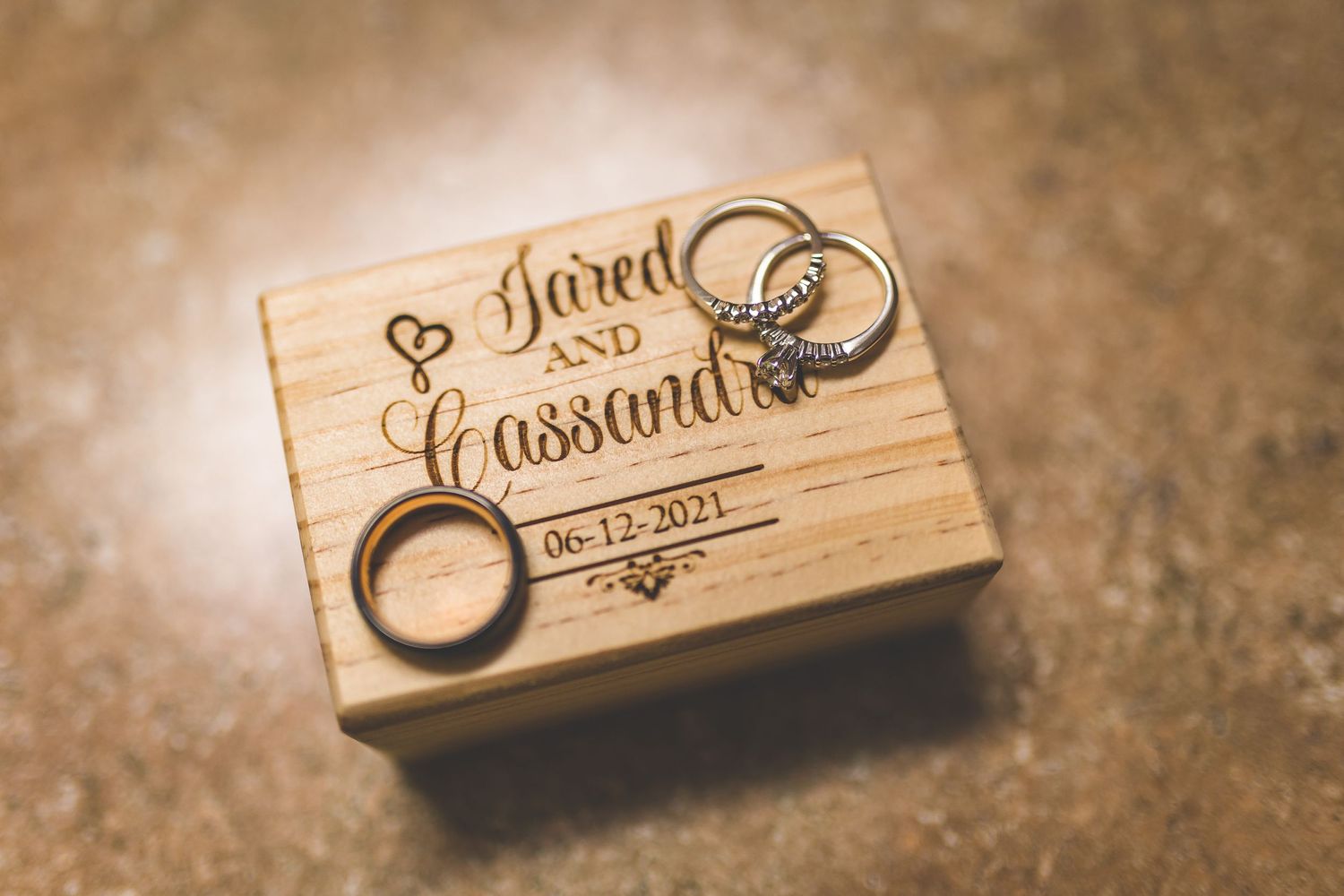 Wedding rings and engraved wooden box with names and date displayed on textured surface.
