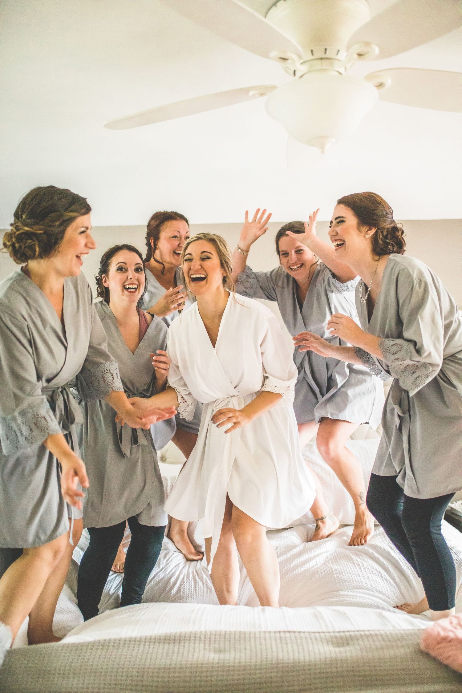 Bridal party in matching robes jumps on bed during pre-wedding preparations.