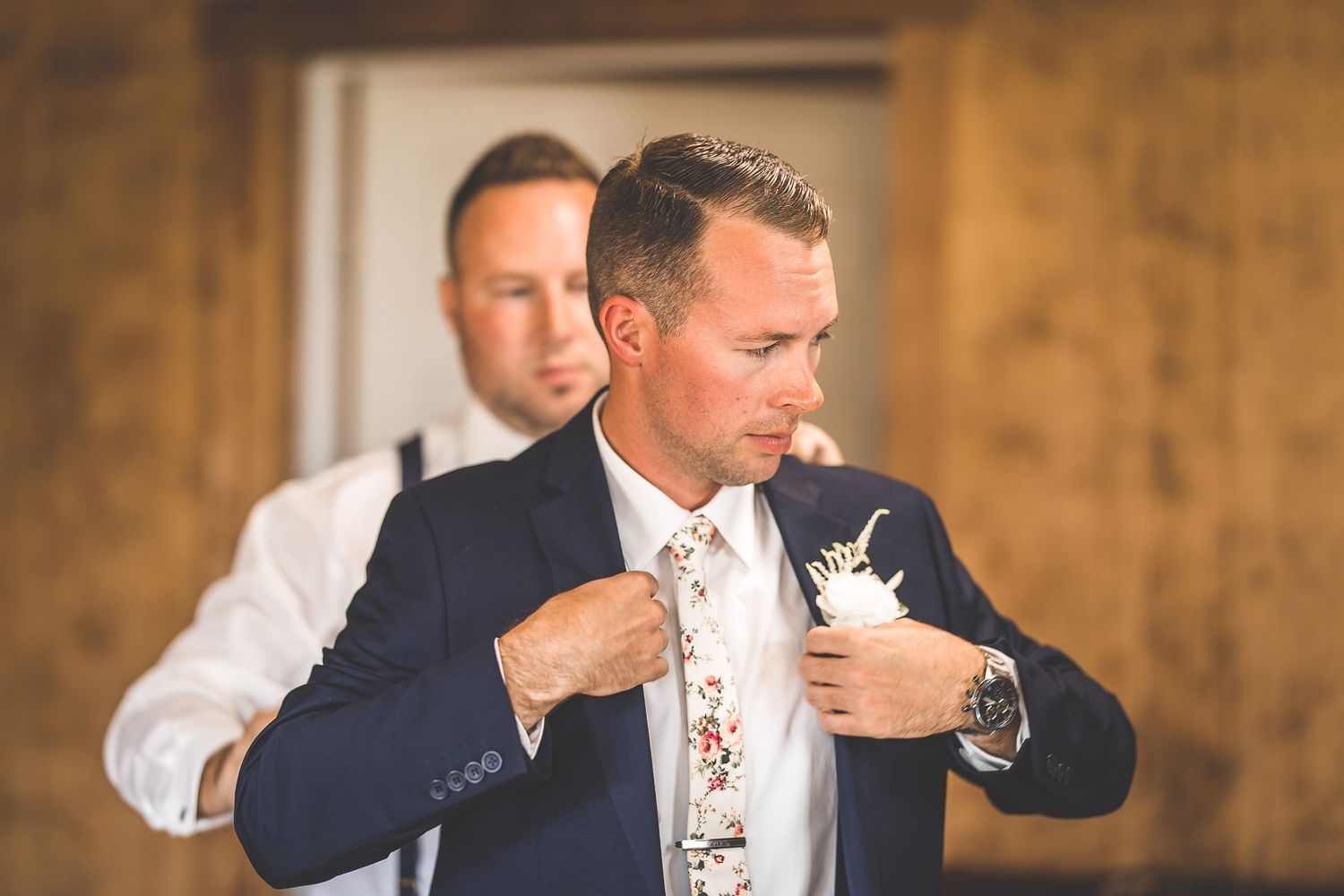 A groom adjusts his tie while getting ready in a navy suit on his wedding day.