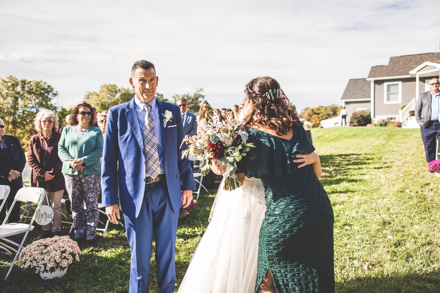 Outdoor wedding ceremony on a grassy hillside with guests seated and a couple exchanging vows under blue skies.