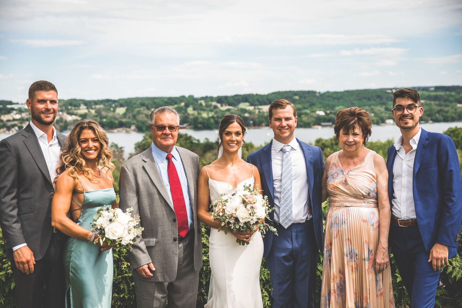 Bridal party members stand together outdoors with lake and hills in background.