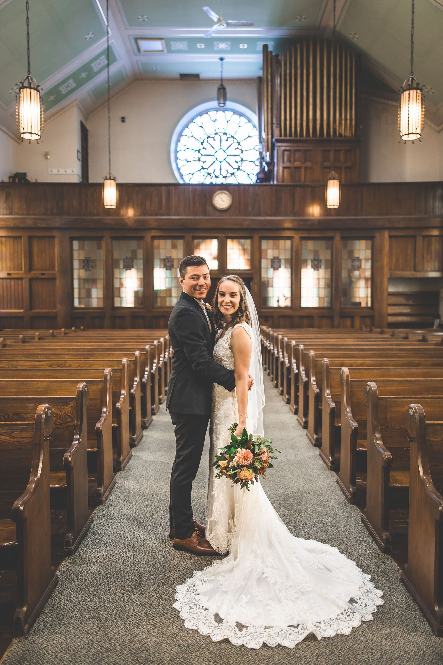 Wedding couple poses in traditional wooden church pews with pendant lighting and stained glass.