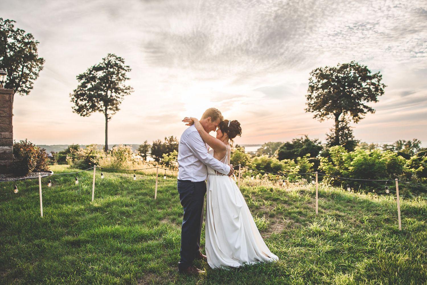 A couple embraces in a scenic field at sunset during their wedding celebration.